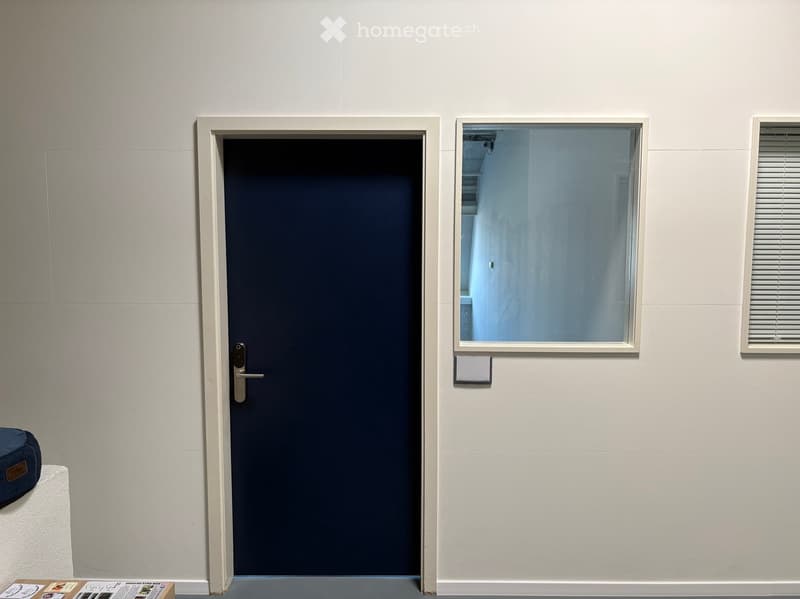 A small, furnished room with a closed dark blue door, a window with blinds, a small table with papers, and a chair.