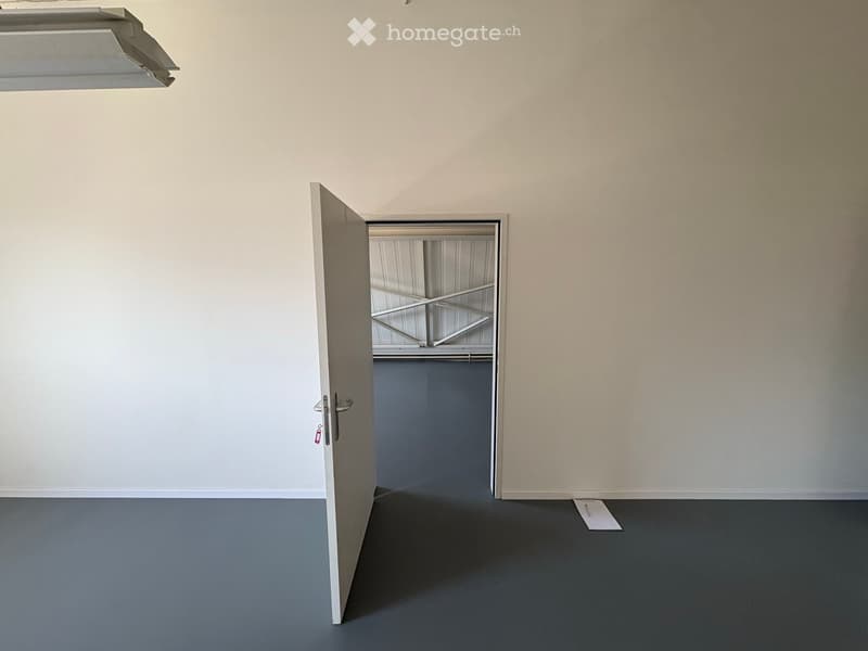 Empty room with grey walls and floor, featuring an open door leading to a storage area with metal shelving.