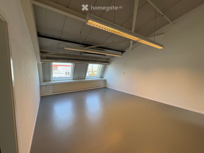 Empty room with white walls, grey floor, exposed ceiling beams, and large windows letting in natural light.