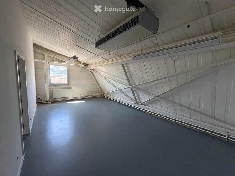 An empty room with a grey floor, white walls, and a large window letting in natural light. The ceiling has exposed beams and a hanging light fixture.