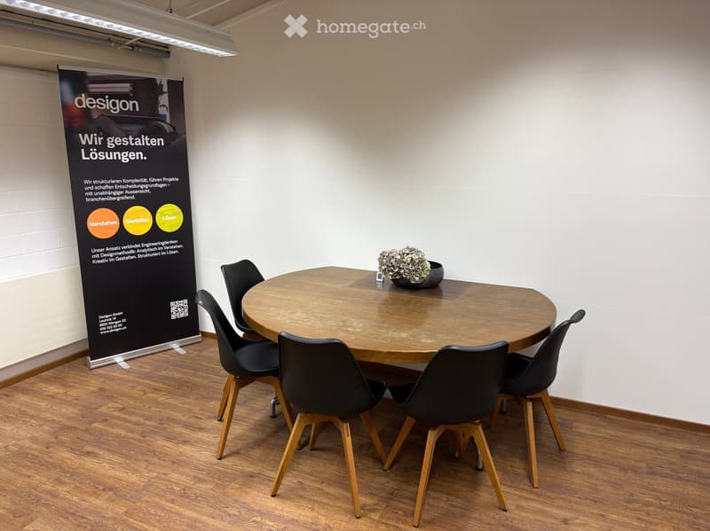A modern meeting room with a round wooden table surrounded by black chairs, a flower pot on the table, and a large banner on the wall.