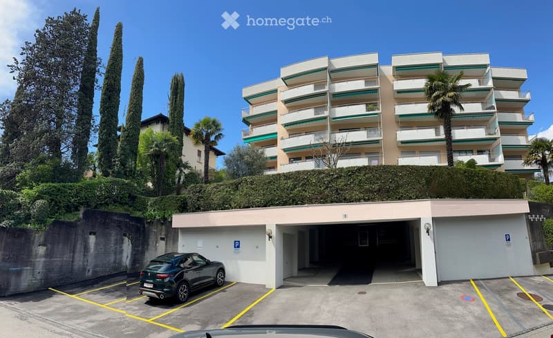 A modern multi-story apartment building with balconies and green awnings, surrounded by tall trees and a hedge. A parking garage is visible in the foreground with a parked car.