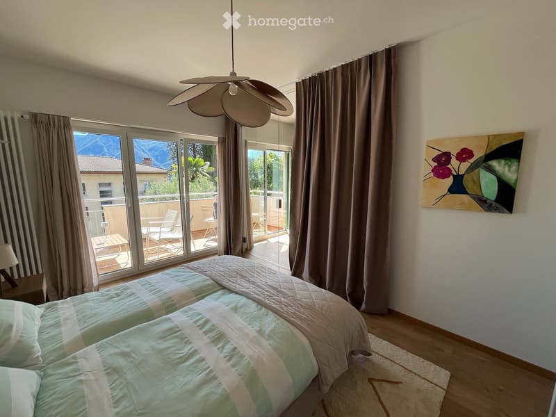 Bright bedroom with two single beds, nightstand, lamp, large window with balcony access, and a modern painting on the wall.