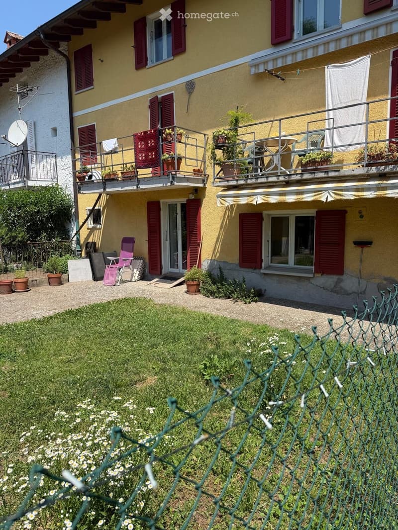 Two-story yellow house with red shutters, balconies, outdoor seating, and garden area.