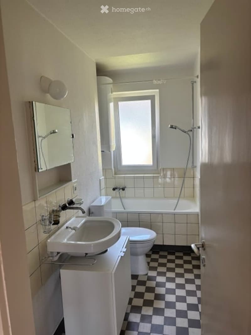 Modern bathroom with white tiles, a sink, mirror, bathtub, toilet, and a window for natural light.