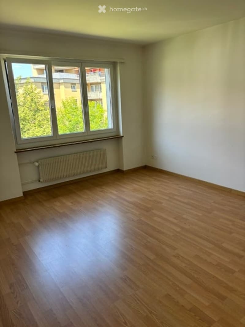 Empty room with wooden flooring, white walls, and a large window with a radiator below it.