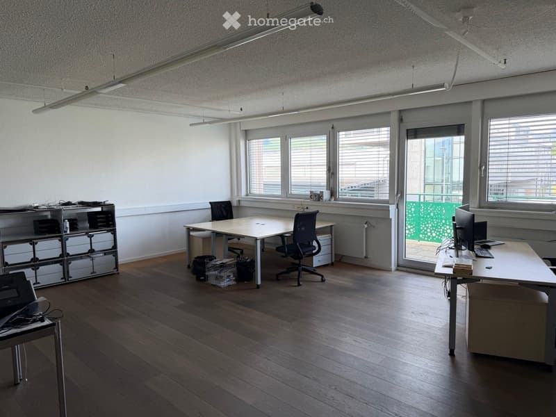 Office space with wooden flooring, large windows, and modern furniture including desks, chairs, and a shelving unit.