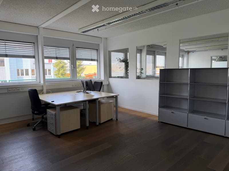 Modern office space with wooden flooring, large windows with blinds, a desk with a computer monitor, office chair, and a metal shelving unit.