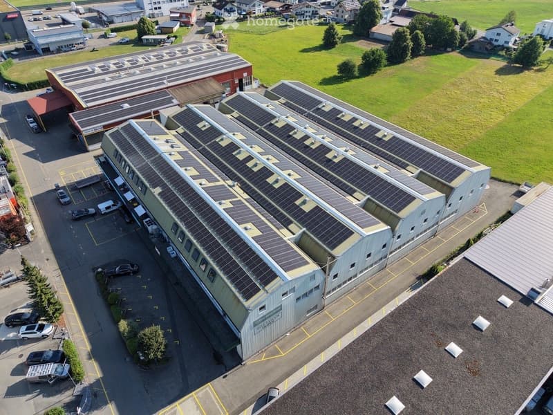 Aerial view of a large industrial building with solar panels on the roof, surrounded by a parking lot and other structures in a rural area.