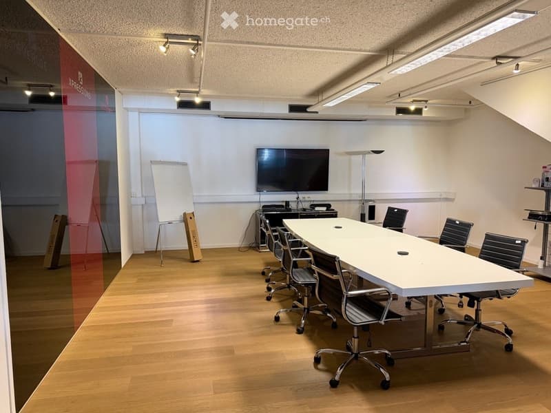 Modern conference room with a long white table, several black chairs, a large screen, a whiteboard, and a computer setup.