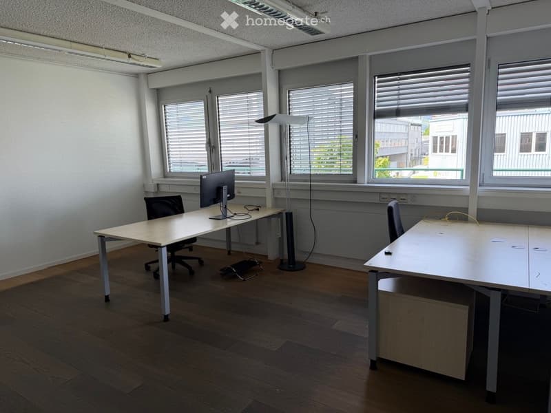 Office space with wooden flooring, two desks, chairs, a computer monitor, and a floor lamp. Large windows with blinds provide natural light.