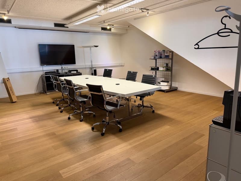 Conference room with a large white table, multiple black chairs, a large screen, a floor lamp, and a small cabinet with various items.