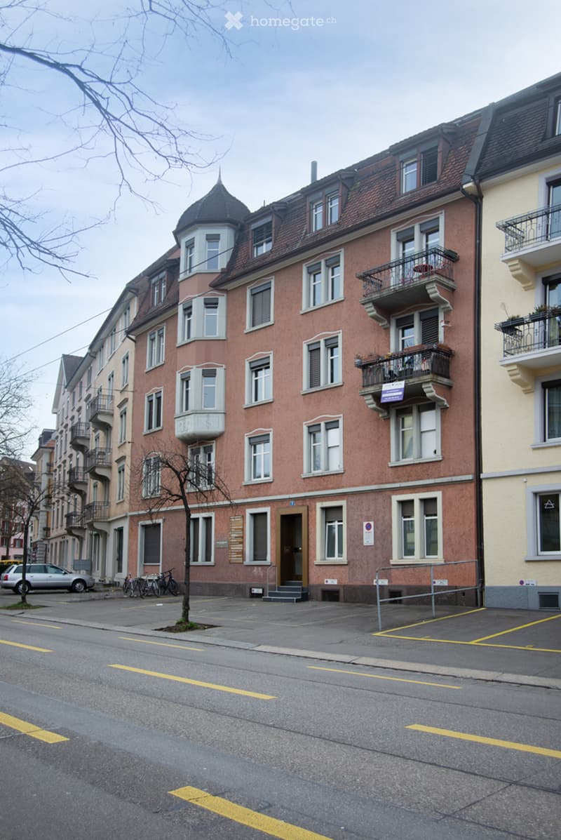 Multi-story residential building with red brick facade, multiple windows, balconies, and a small tree in front.