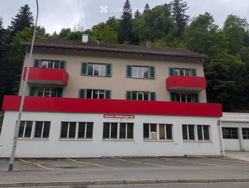 A multi-story building with a beige facade, green shutters, and red balconies. The ground floor has large windows and a sign reading "Eduard Waldburger AG".