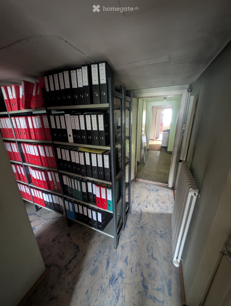 A narrow hallway with a metal shelving unit filled with binders and files, a radiator on the right wall, and a visible room at the end.