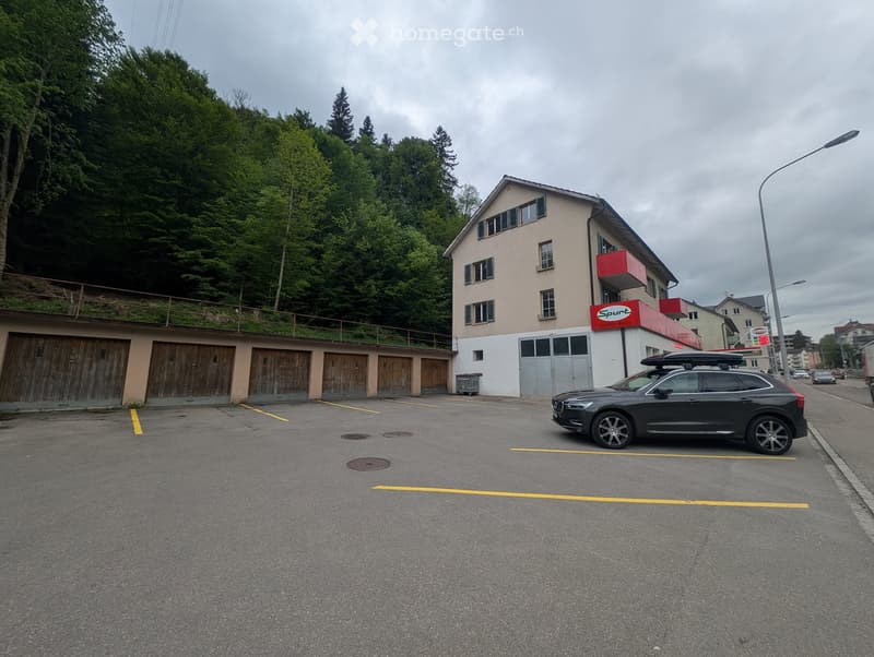A multi-story building with a garage area, a parked car, and a red sign on the roof. The building is surrounded by greenery and has a modern design.