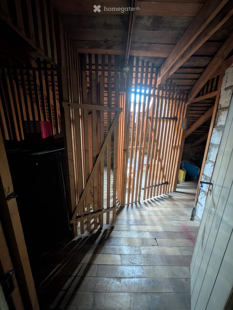 An unfinished attic corridor with exposed wooden beams and a staircase leading upwards, featuring a wooden railing and tiled floor.
