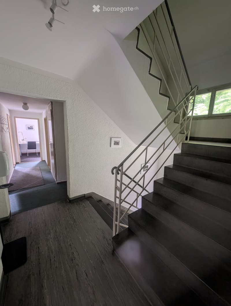 Staircase with wooden steps and metal railing, leading to an open area with a visible room on the left side. The hallway has light-colored walls and a wooden floor.