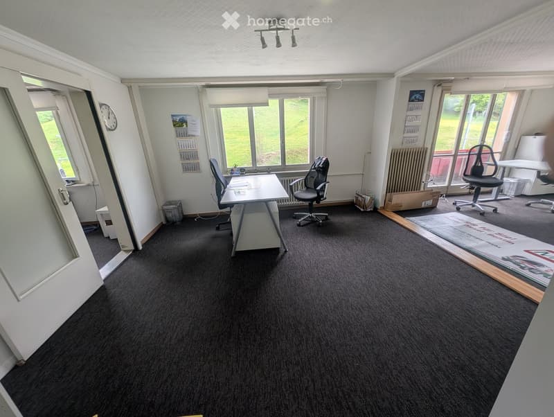 Office space with carpeted floor, large windows, desks, chairs, and a calendar on the wall.