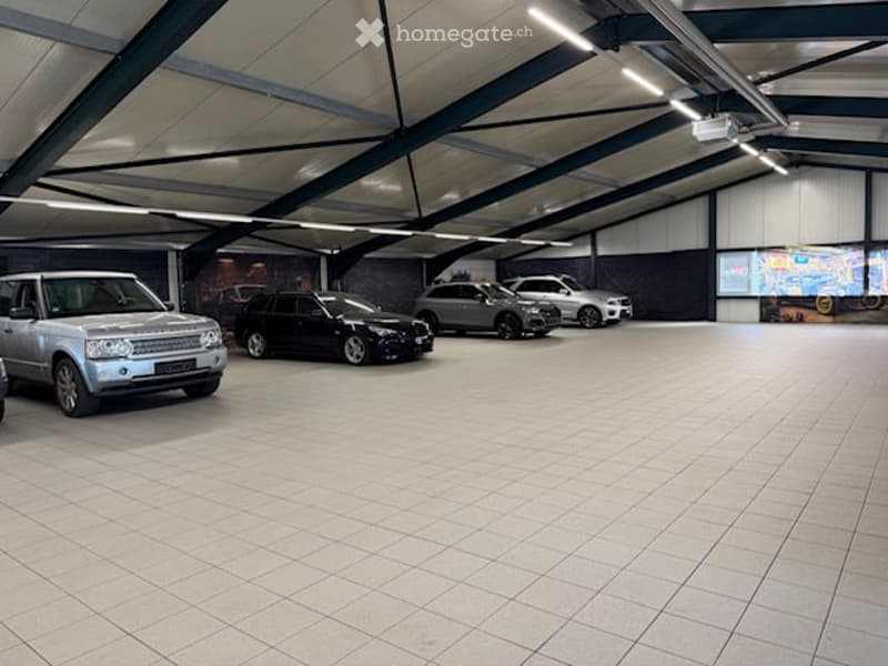 Spacious indoor parking area with multiple parked cars, tiled floor, and high ceiling with exposed beams and lighting.