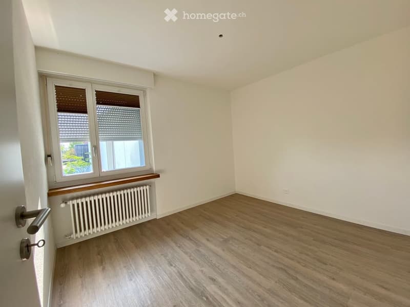 Empty room with light-colored walls, wooden flooring, a window with blinds, and a radiator below the window.