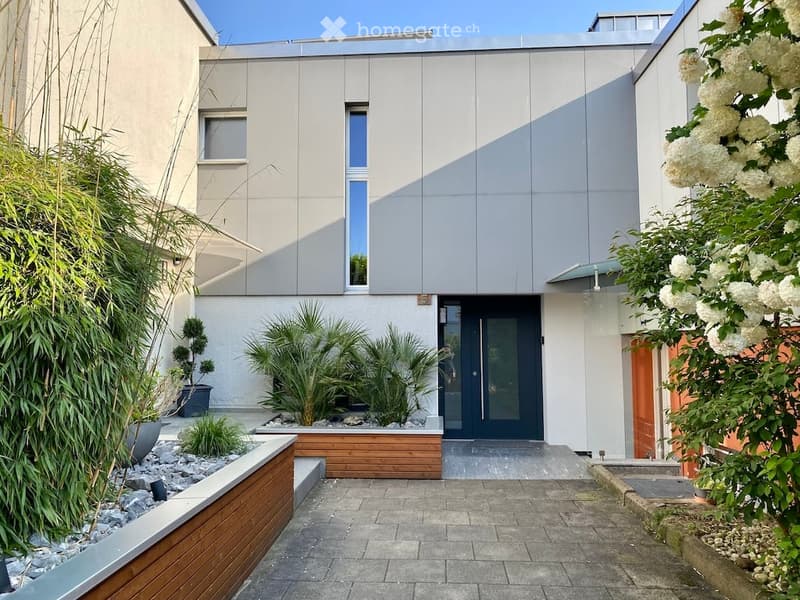 Modern two-story house with a sleek, contemporary design featuring a combination of grey and white exterior walls, large windows, and a dark front door. The entrance is flanked by landscaped garden areas with bamboo, palm plants, and neatly arranged rocks