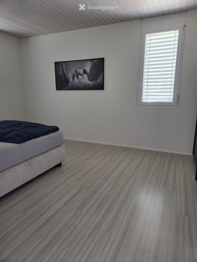 Bedroom with a grey bed, dark blue blanket, and a picture frame on the wall. The room has light grey walls and a grey, textured wooden floor.