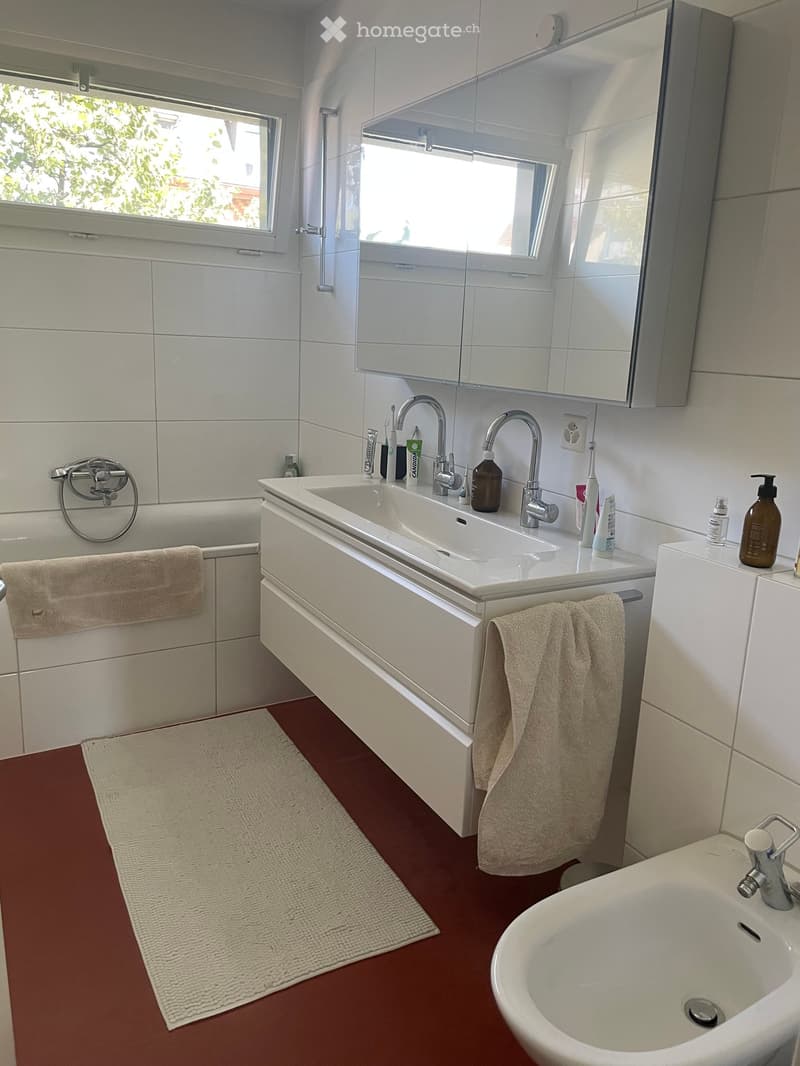 Modern bathroom with white tiles, a double sink, a bathtub, and a large mirror. The floor is red, and towels are placed on the sides.