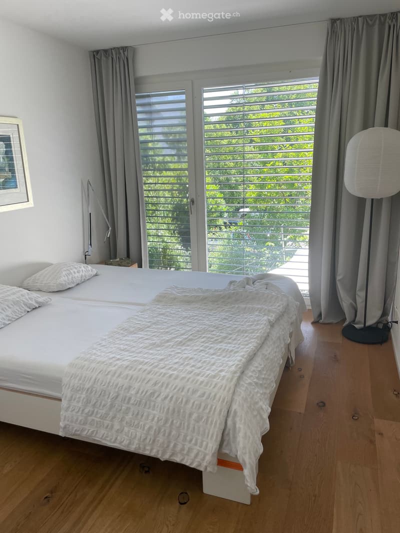 A modern bedroom with a large bed, white bedding, and a textured blanket. The room features wooden flooring, large windows with blinds, and a floor lamp near the window.