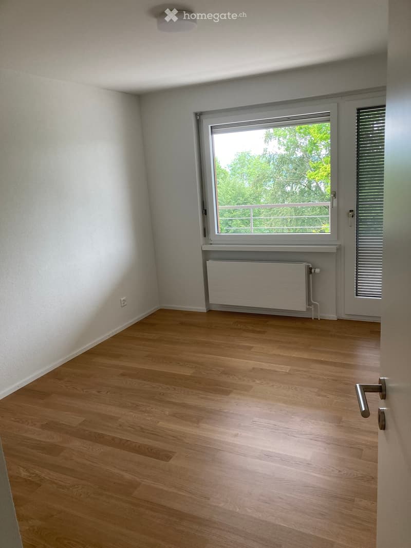 Empty room with wooden flooring, white walls, a window with a view of greenery, a radiator, and a door with a handle.