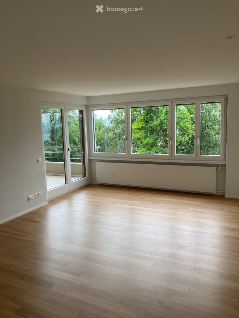 Empty room with wooden flooring, large windows, and a radiator. Access to a balcony or terrace is visible through a sliding glass door.