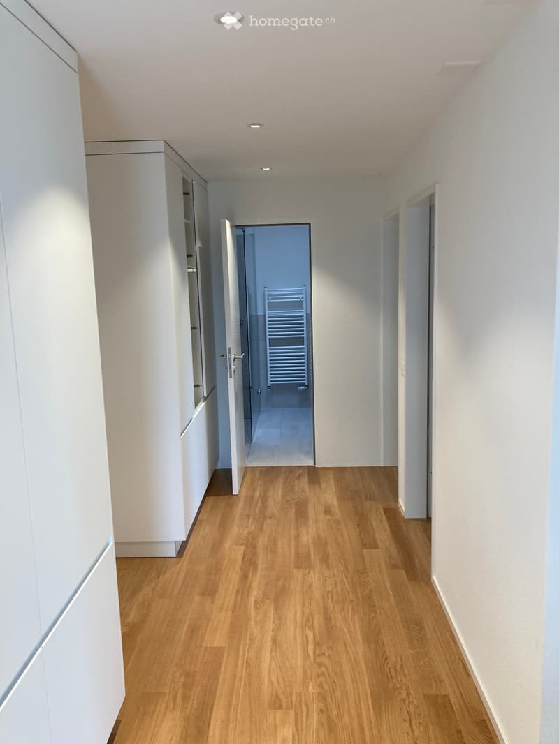 A modern, minimalist hallway with light wooden flooring, white walls, and recessed ceiling lighting. An open door leads to a tiled bathroom with a radiator visible.