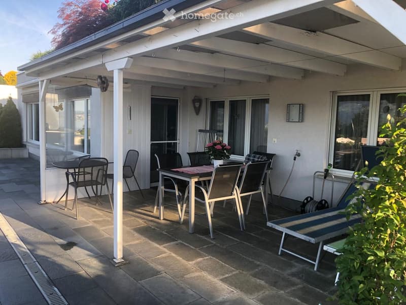 Covered patio with a table, chairs, and a lounge chair, surrounded by large windows and a view of the garden.
