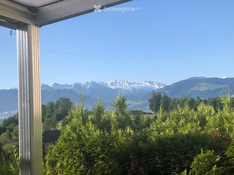 Balcony with a view of mountains and greenery, featuring a metal railing and a clear blue sky.