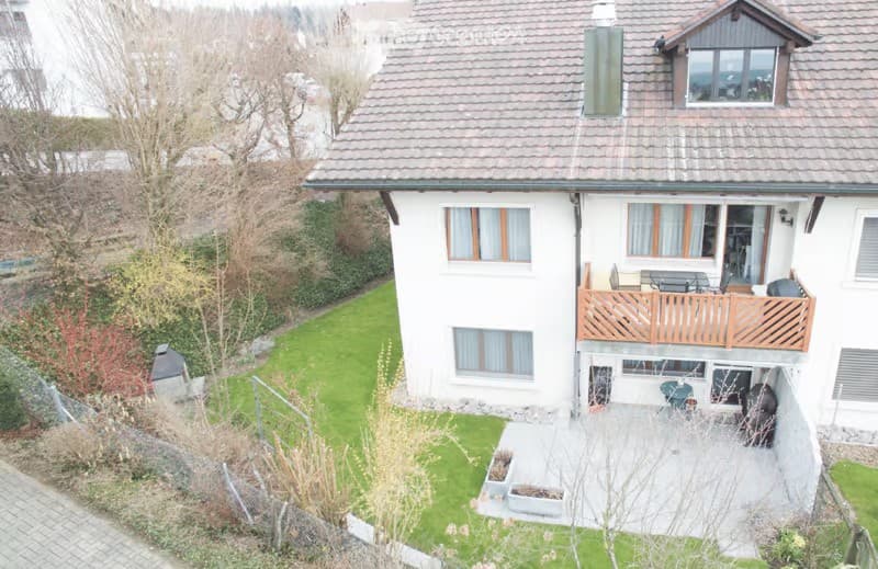 A two-story house with a tiled roof, white exterior, and multiple windows. The house features a wooden balcony with outdoor furniture and a well-maintained lawn.