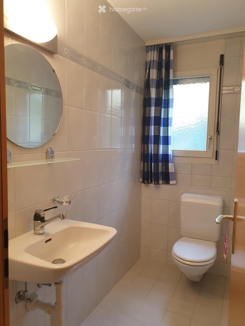 A small bathroom with a white sink, toilet, round mirror, and a window with a blue and white checkered curtain.