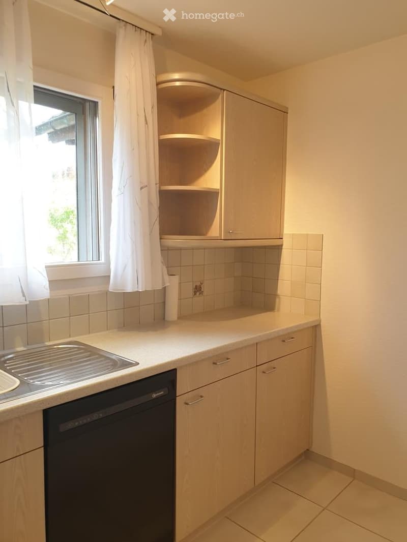 Modern kitchen with light wooden cabinets, white countertops, a sink, and a dishwasher. A window with a curtain is visible on the left side.