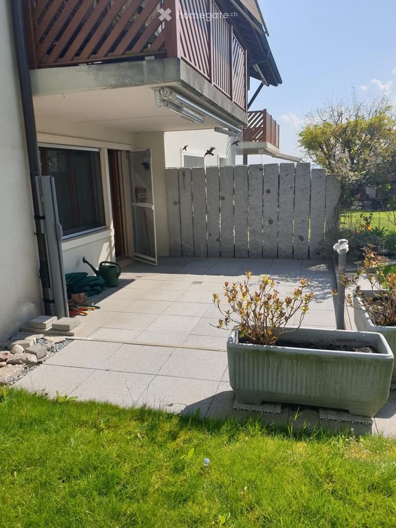 Balcony area with tiled floor, concrete planter, small garden, and adjacent house with balcony and wooden fence.