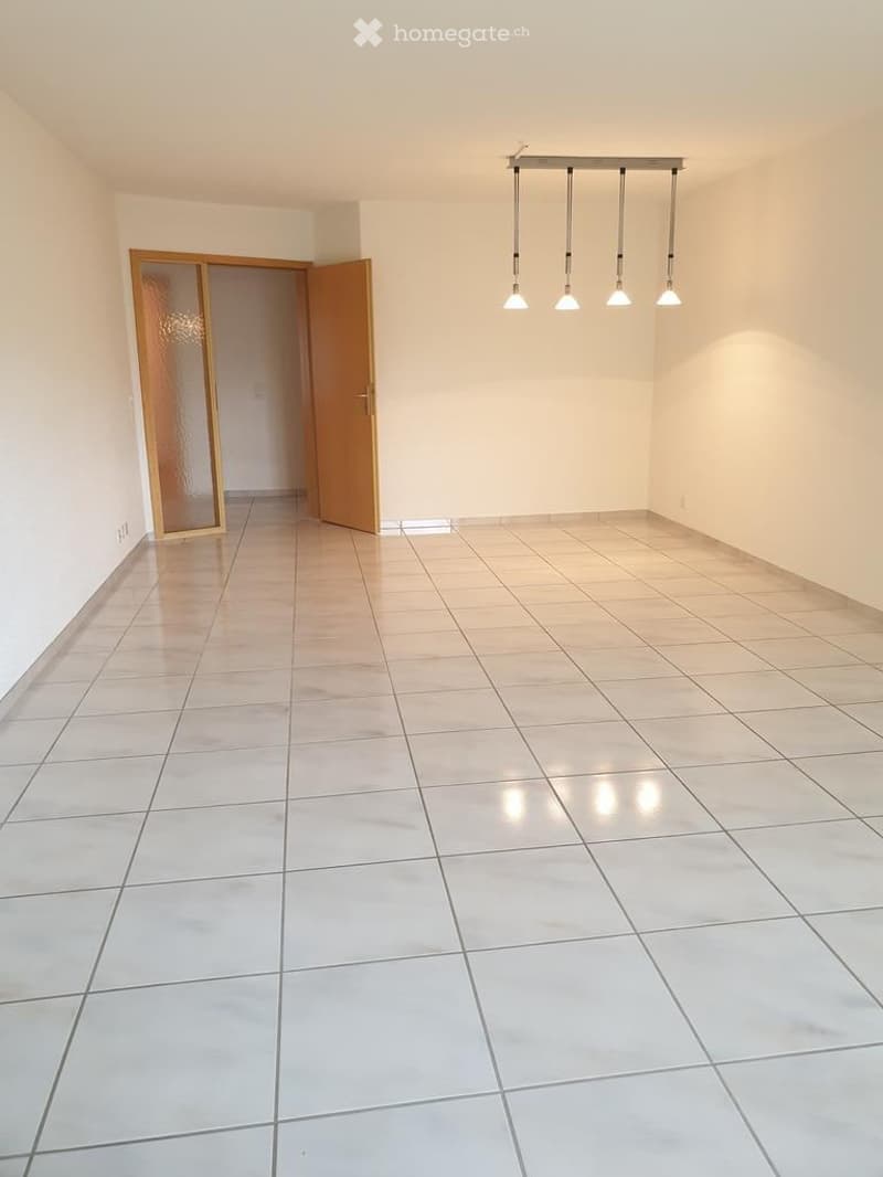 Empty room with white tiled flooring, light-colored walls, and a wooden door leading to another space. A modern pendant light fixture hangs from the ceiling.
