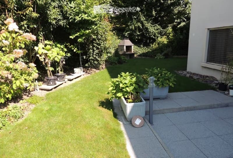 Well-maintained garden with a neatly trimmed lawn, several potted plants, a small barbecue area, and a paved patio area.