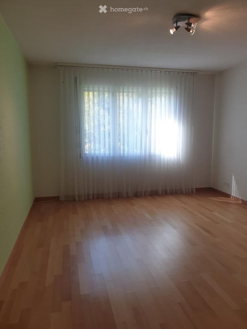 Empty room with wooden flooring, large window with white curtains, and a ceiling light fixture.