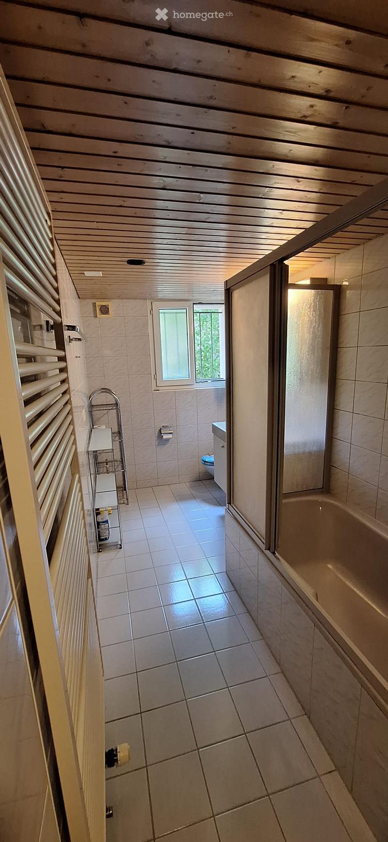Bathroom with tiled floor and walls, a bathtub, toilet, radiator, and a window with blinds.