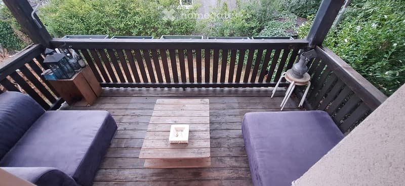 Wooden balcony with two purple cushions, a small wooden coffee table, and a small stool with a decorative item on it.
