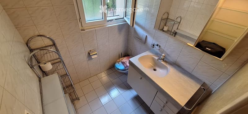 Modern bathroom with white tiles, a sink with a faucet, a toilet, a mirror, and a towel rack. The room has a window with a view of greenery outside.