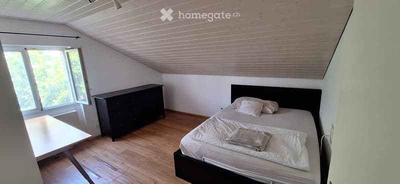 Bright bedroom with wooden flooring, slanted wooden ceiling, large window with open shutters, bed with white bedding, nightstand, dresser, and small table.