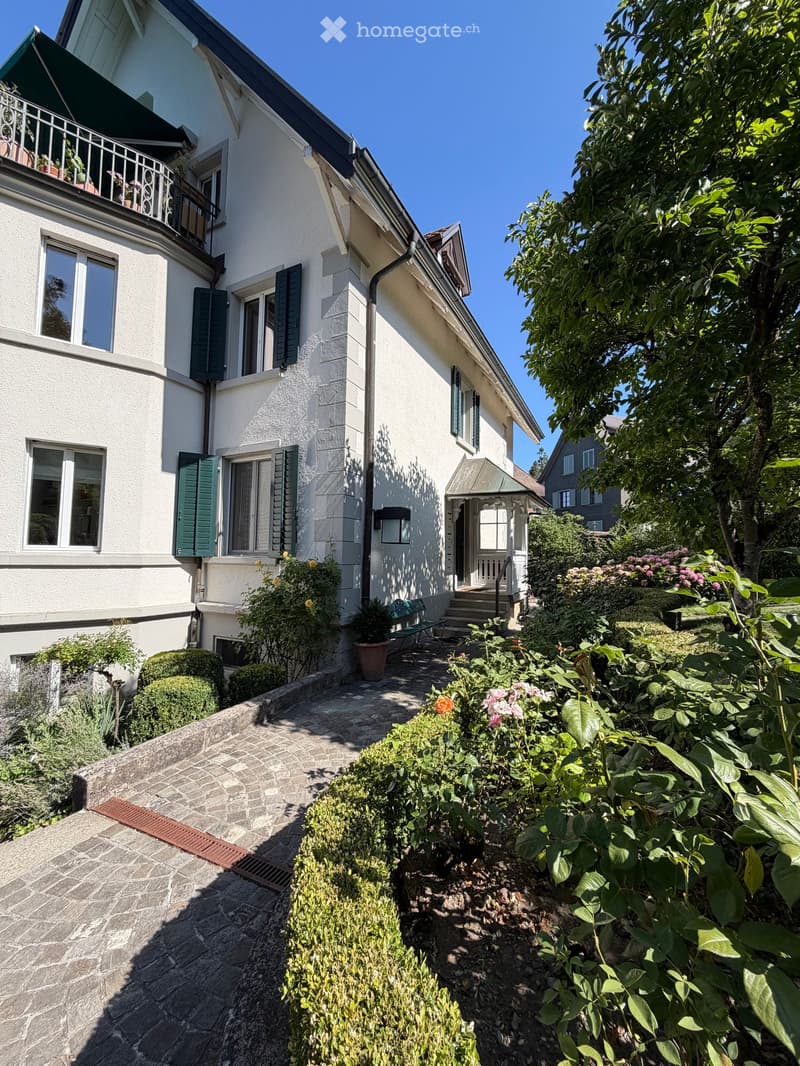 Two-story white house with green shutters, balcony, and well-maintained garden with flowers and shrubs.