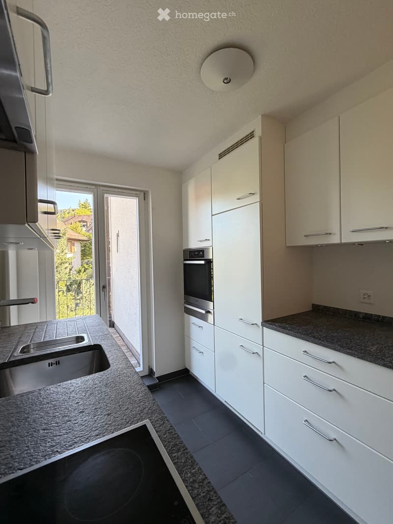 Modern kitchen with white cabinets, granite countertops, stainless steel sink, oven, and stove. Features a large window with a view of greenery.