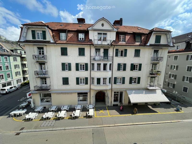 4-story residential building, white exterior, green shutters, red roof, multiple balconies, outdoor seating with white chairs and tables, awning over entrance, parking space in front, visible views of other buildings and street