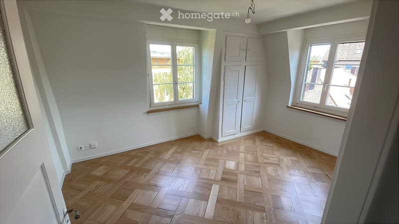 Empty room with wooden herringbone flooring, white walls, built-in white closet, and two windows with wooden sills.