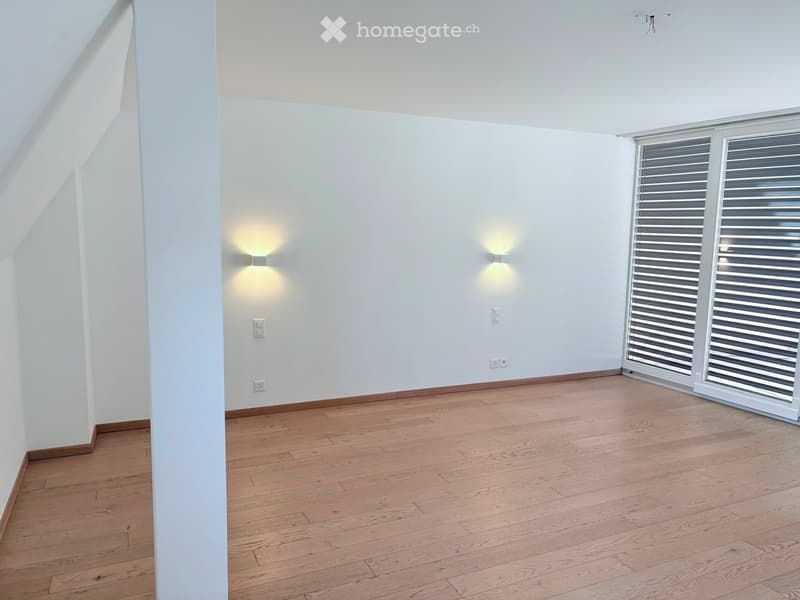 Empty room with light wooden flooring, white walls, and large windows with blinds. Minimalist design with wall-mounted lighting and electrical outlets.
